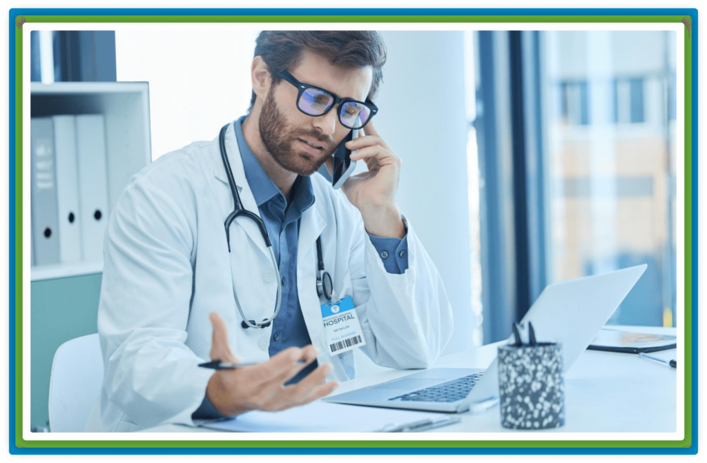 A doctor in glasses and a white coat talks on phone, using a laptop.